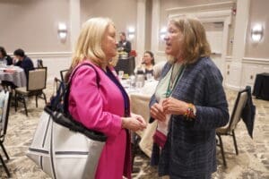 Two women stand and talk in a conference room with tables and other people seated in the background.