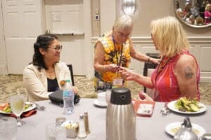Three women sit at a round table with plates of food and drinks, engaging in conversation at an indoor event or conference.