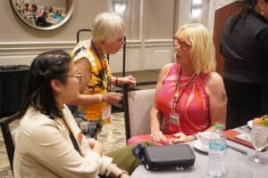 Three people in conversation around a table at an indoor event, with conference badges visible; one person is standing while two are seated.