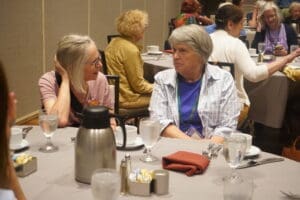 Two older women sit at a round table in a restaurant or event, conversing. There are dishes, water glasses, and a coffee pot on the table. Other people are seated in the background.