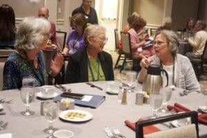 Three elderly women sit at a round table, engaged in conversation during a conference meal, with others seated at tables in the background.