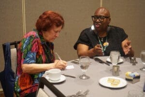 Two women sit at a table; one writes in a notebook while the other gestures with her hands. There are drinks, dishes, and snacks on the table.