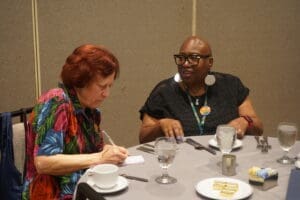 Two women sit at a round table with place settings; one writes in a notebook while the other, wearing glasses and earrings, talks and gestures with her hands.