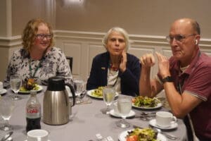Three people sit at a round table eating salad, with glasses of water, a Diet Coke can, and a metal coffee pot in the center.