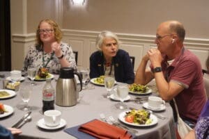 Three adults sit around a round table with plates of salad, drinks, and cutlery, engaged in conversation at what appears to be a formal meal or meeting.