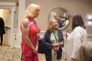 Three women are standing together in conversation at an indoor event; one wears a bright pink dress, and two have attendee lanyards. A round mirror and people are visible in the background.