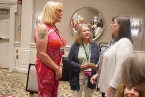 Three women are standing and conversing in a conference room; one woman wears a bright dress with a visible arm tattoo, and a round mirror is on the wall behind them.