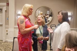 Three women are standing and talking in a conference room. One woman is holding a piece of paper while another wears a bright pink dress. A round mirror hangs on the wall behind them.