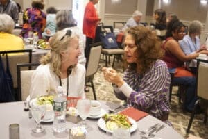 Two women sit at a round table with salads and drinks, engaged in conversation at a crowded indoor event or luncheon.