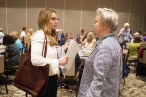 Two women stand and talk at a crowded event or conference, with people sitting and eating at tables in the background.