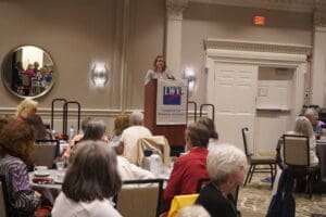 A woman speaks at a podium labeled “League of Women Voters” to an audience seated at round tables in a conference room.