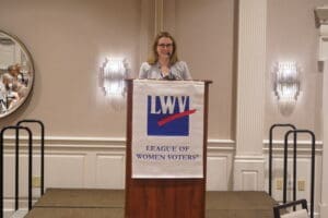 A woman stands at a podium with a "League of Women Voters" banner, speaking to an audience in a conference room.