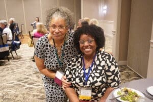 Two women wearing name badges and patterned dresses pose and smile at a round table during a conference or event. Other attendees are seated in the background.