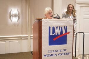 An older woman speaks at a podium with an LWV (League of Women Voters) banner, while another woman stands in the background.