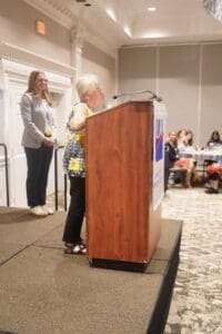 An older woman speaks at a podium in a conference room while another woman stands nearby and attendees sit at tables in the background.