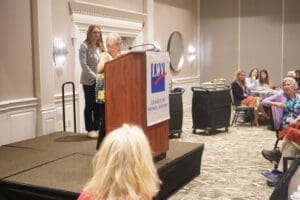A woman is speaking at a podium with a "League of Women Voters" sign in a conference room while attendees listen and watch.