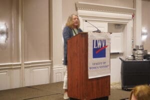 A woman speaks at a podium with an LWV (League of Women Voters) banner in a conference room.