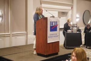 A woman speaks at a podium with a "League of Women Voters" banner in a conference room; people work in the background.
