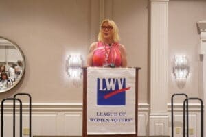 A woman wearing a pink dress speaks at a podium with a "League of Women Voters" banner in a formal event setting.