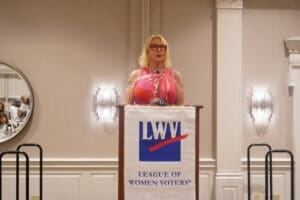 A person wearing a pink dress stands at a podium with an "LWV League of Women Voters" banner in a conference room.