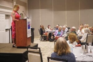 A person stands at a podium addressing a seated audience in a conference room. The podium has a sign with "LWV" and "League of Women Voters.