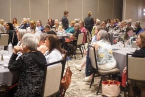A large group of people sit at round tables in a conference room, listening attentively to an unseen speaker.