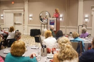A woman speaks at a podium with an LWV sign in front of an audience seated at round tables in a conference room.