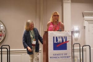 Two women stand at a podium with an LWV (League of Women Voters) banner; one woman speaks into the microphone while the other stands beside her.