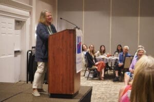 A woman stands at a podium giving a speech to an audience of seated women in a conference room.