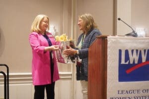 Two women stand together as one hands a gift bag to the other at a podium with a "League of Women Voters" banner in a conference room.