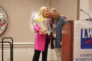 Two women stand together at a podium, one holding a wrapped gift and flowers, in a conference room with a "League of Women Voters" banner.
