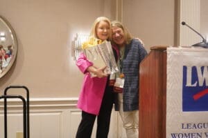 Two women stand together smiling at an event; one holds a gift bag. A podium with an "LWV" banner is visible on the right.