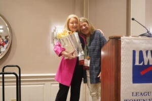 Two women stand together and smile; one holds a gift bag with tissue paper. They are beside a podium with a League of Women Voters banner in a conference room.