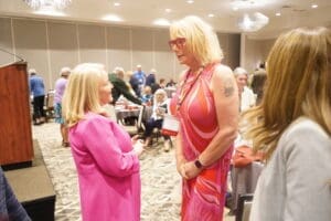 Two women in conversation at a formal indoor event, both wearing pink clothing, with other attendees seated at tables in the background.