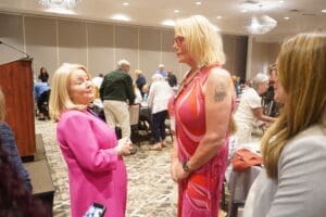 Two women stand and talk in a crowded conference room with round tables and people seated and mingling in the background.