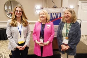 Three women stand and smile in front of a podium with a "LWV" banner at a formal indoor event.