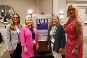 Four women stand in front of a podium with a "League of Women Voters" banner inside a conference room, posing for a group photo.