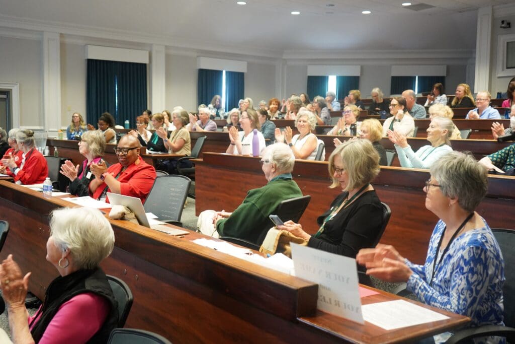 A group of adults seated in a lecture hall clap and listen to a presentation, with some taking notes and others using electronic devices.
