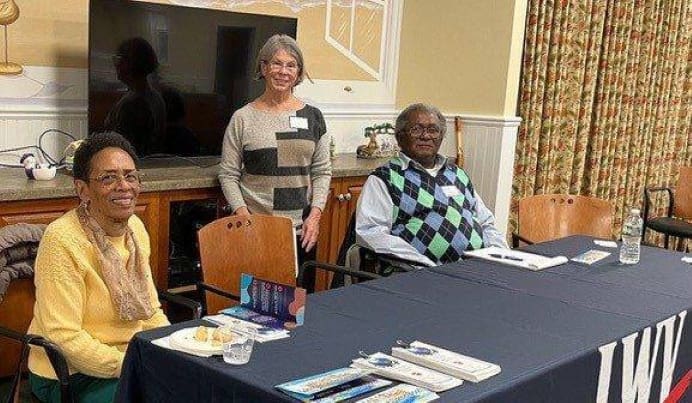 Three adults sit and stand behind a table with books, water, and snacks in a conference room setting.