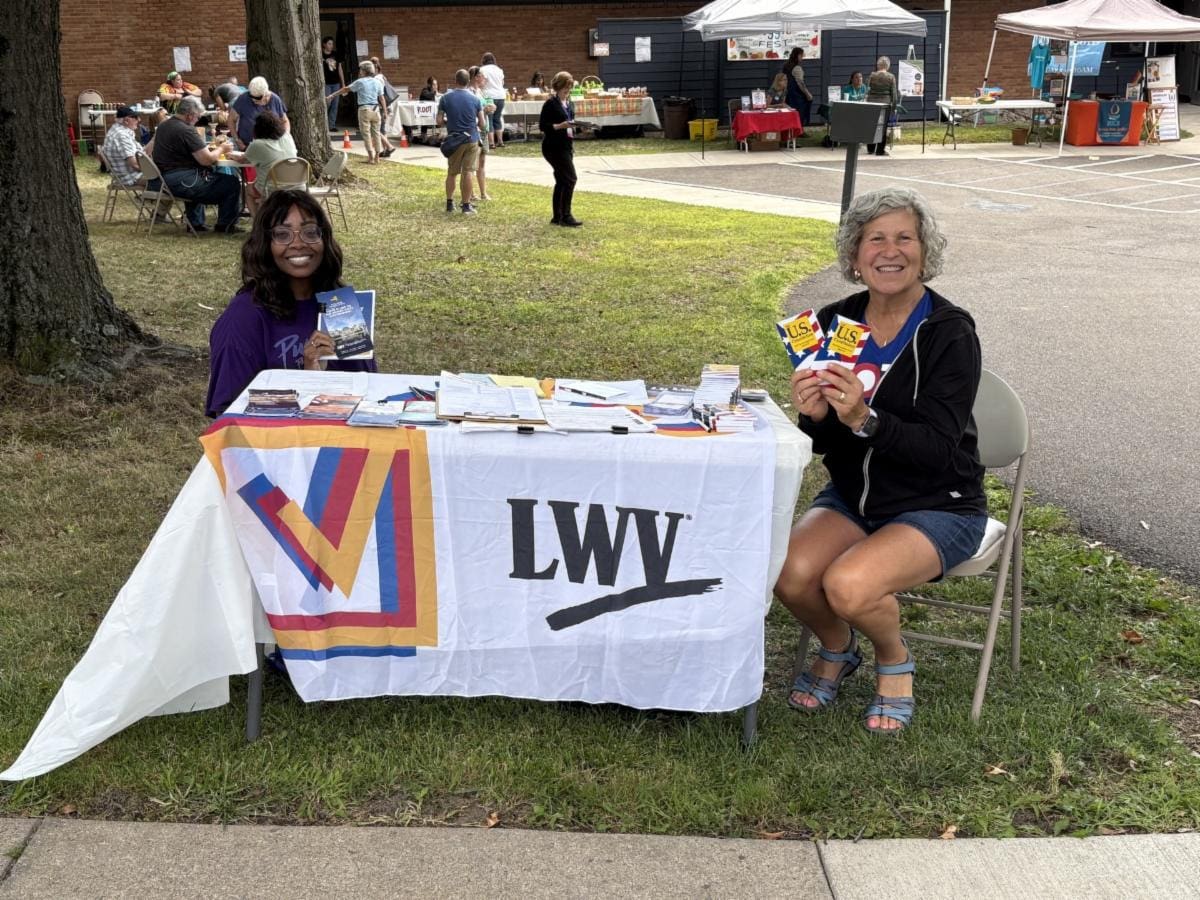 Two women sit at an outdoor table with an LWV banner, holding pamphlets. Other people and vendor booths are visible in the background.