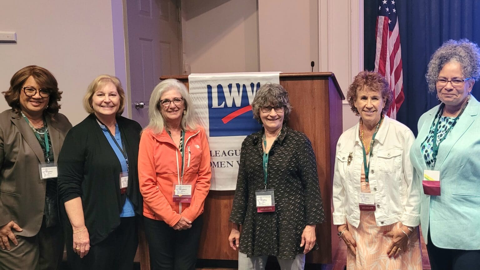Six women stand in front of a podium with a "League of Women Voters" banner, posing for a group photo in a conference room.