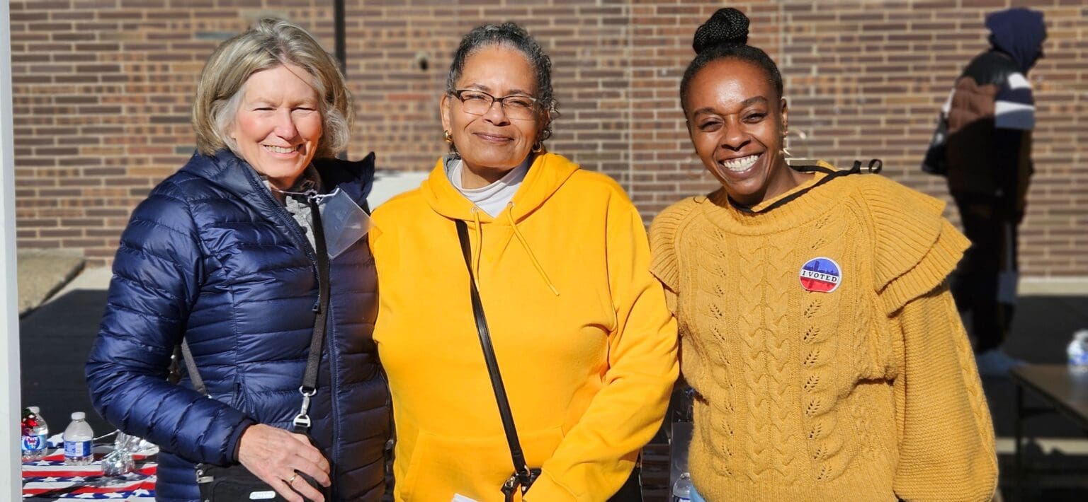 Three women stand outdoors smiling at the camera; two wear yellow sweaters and one wears a blue jacket. A political sticker is visible on one woman's sweater.