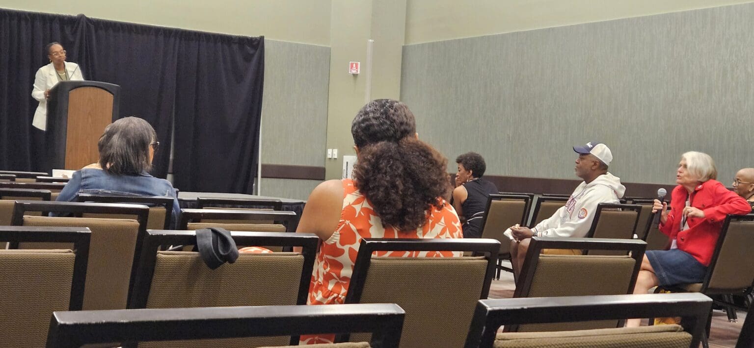 A speaker stands at a podium addressing a small seated audience in a conference room.