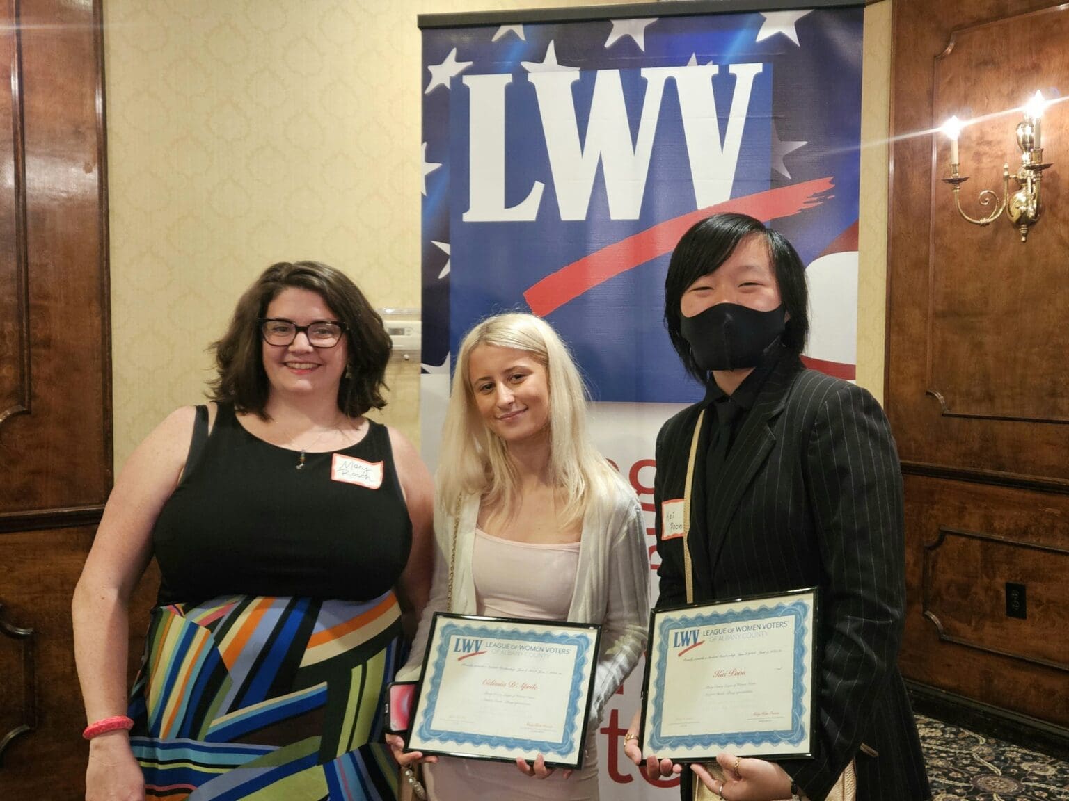 Three people stand indoors in front of an LWV banner; two hold certificates and one is wearing a face mask.